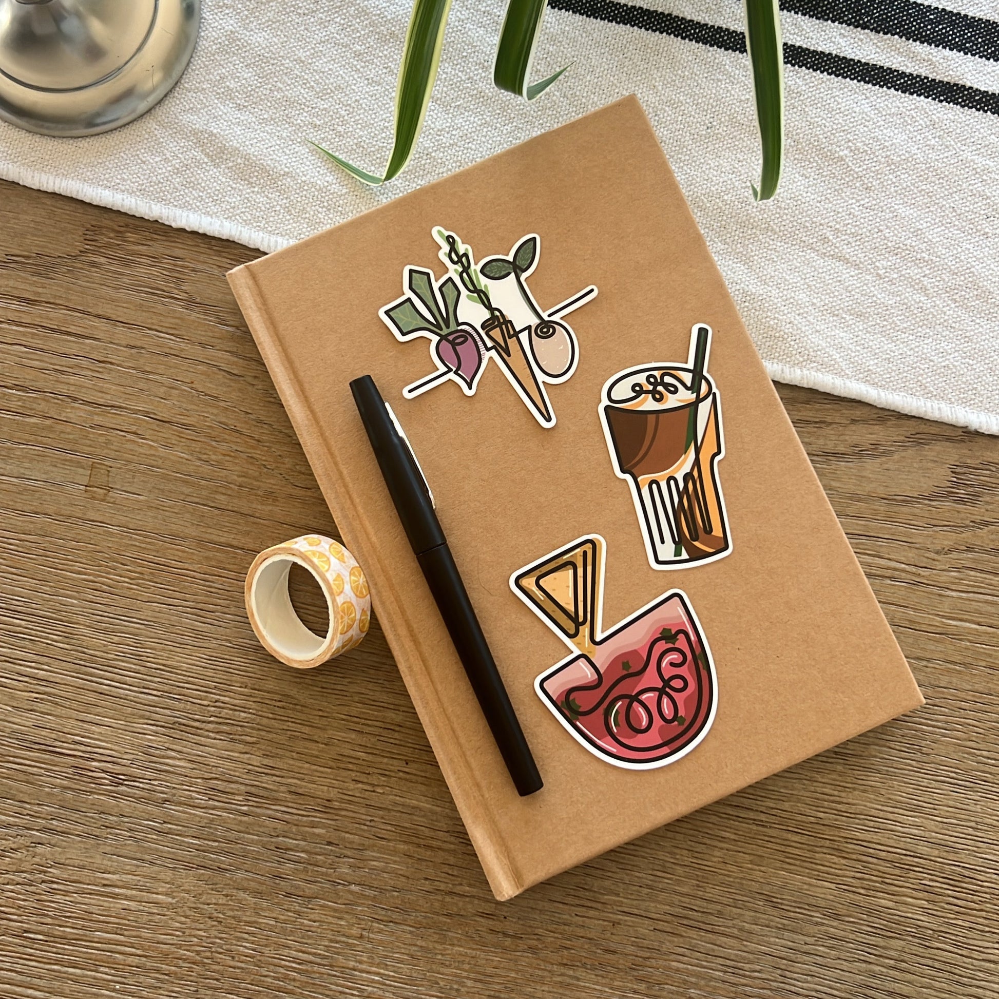 Notebook with stickers on a wooden surface, next to a pen and a plant