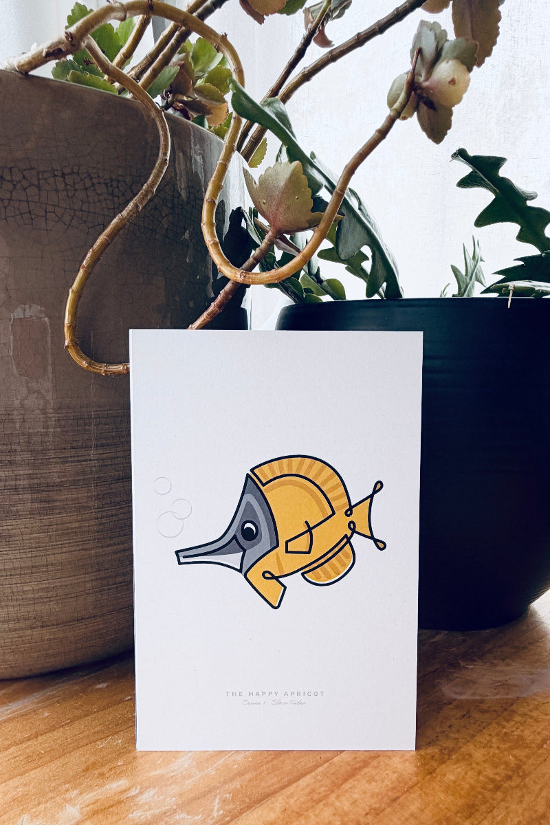 A white greeting card featuring a minimalist cartoon seahorse illustration in yellow, gray, and black, standing on a wooden surface with potted plants in the background.