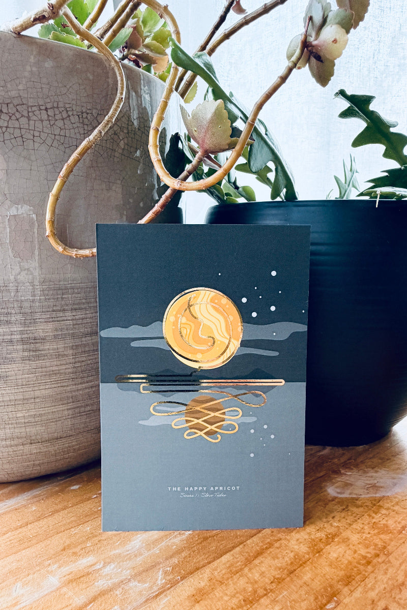 A greeting card with a dark gray background and gold foil illustration of a full moon and reflection, standing on a wooden surface between potted plants.