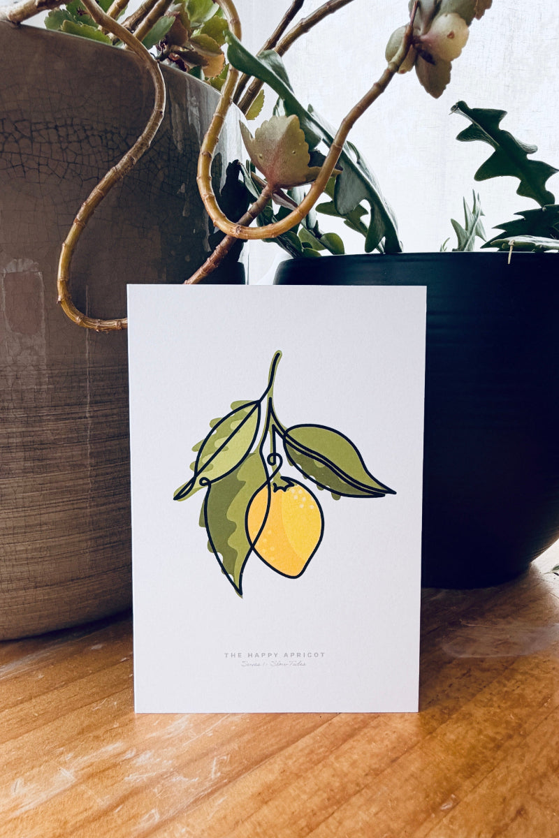 A white greeting card featuring a minimalist illustration of a yellow lemon with green leaves, standing on a wooden table in front of potted plants.