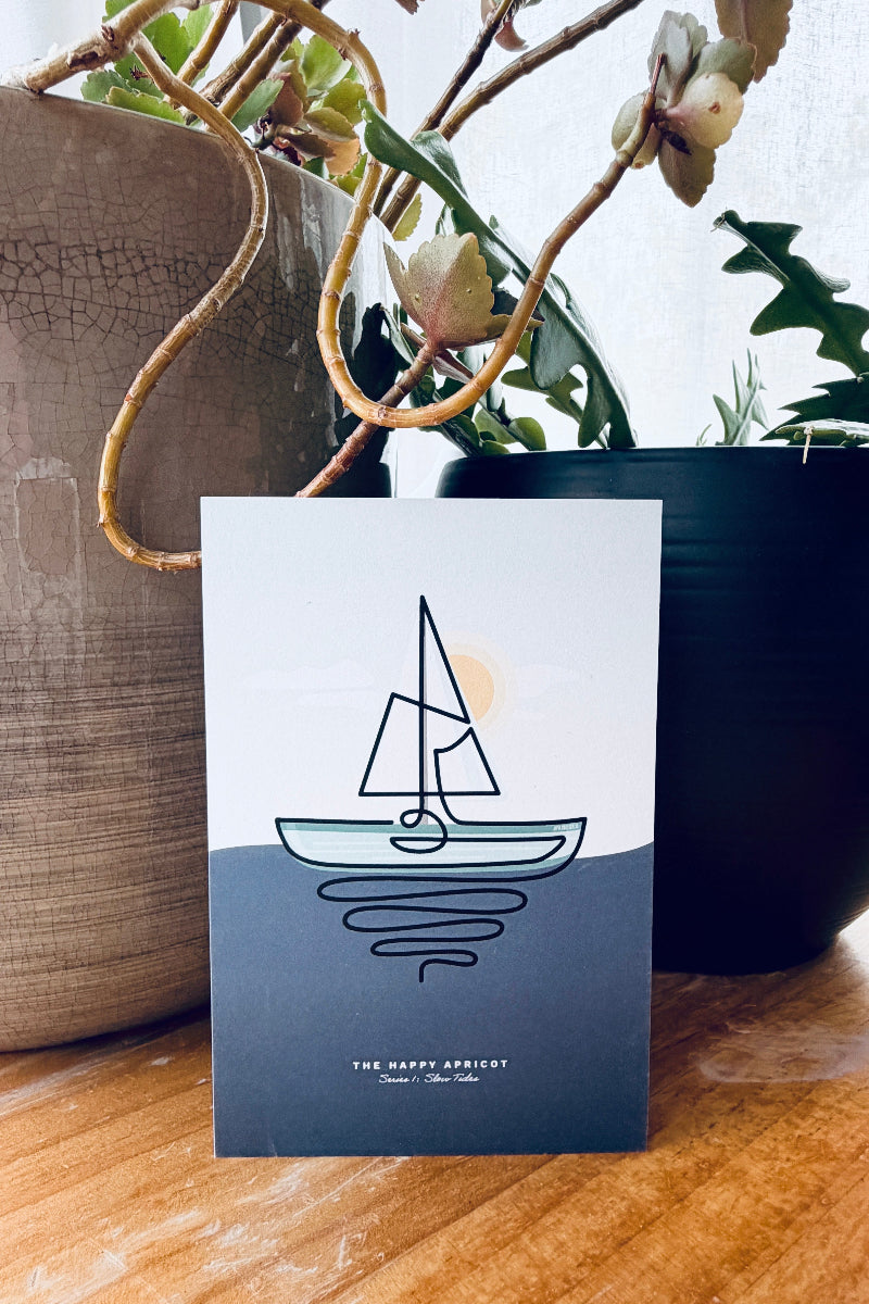 A minimalist art print card titled 'THE SKY IS THE SEA' featuring a simple line drawing of a sailboat on a split white and dark blue background, displayed on a wooden table between two potted plants.