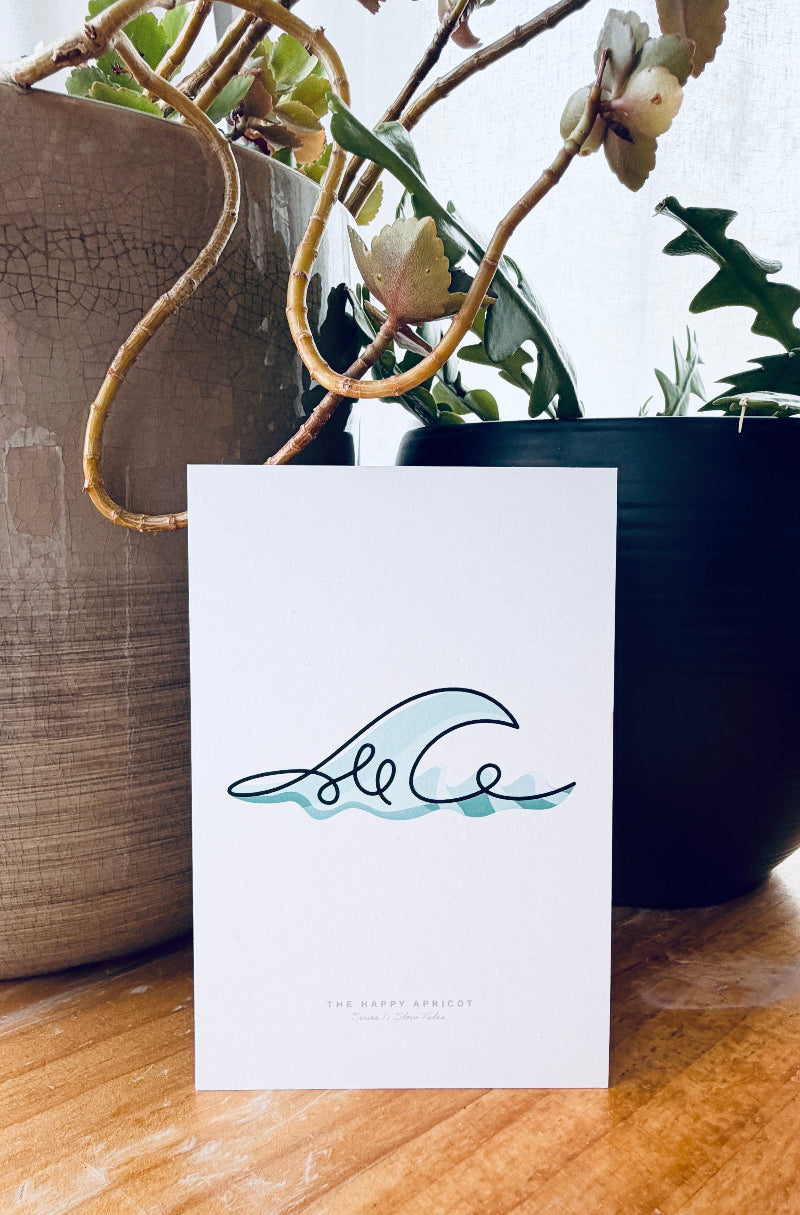 A white art print card featuring a minimalist black and teal wave illustration, displayed on a wooden surface with potted plants in the background.