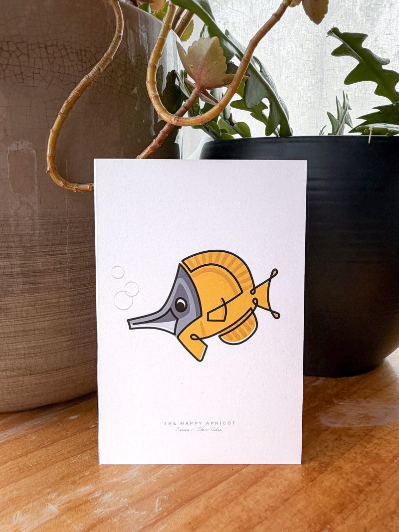 A white greeting card featuring a cartoon yellow apricot fish with a gray face, standing on a wooden surface in front of potted plants.