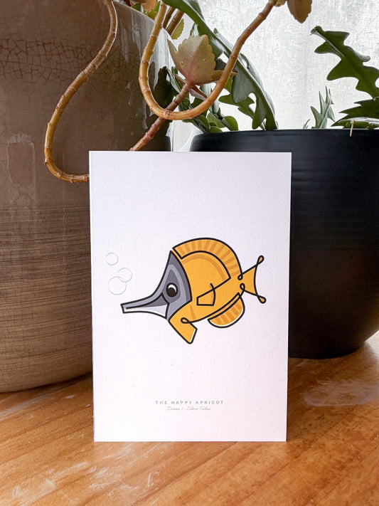 A white greeting card featuring a cartoon yellow apricot fish with a gray face, standing on a wooden surface in front of potted plants.