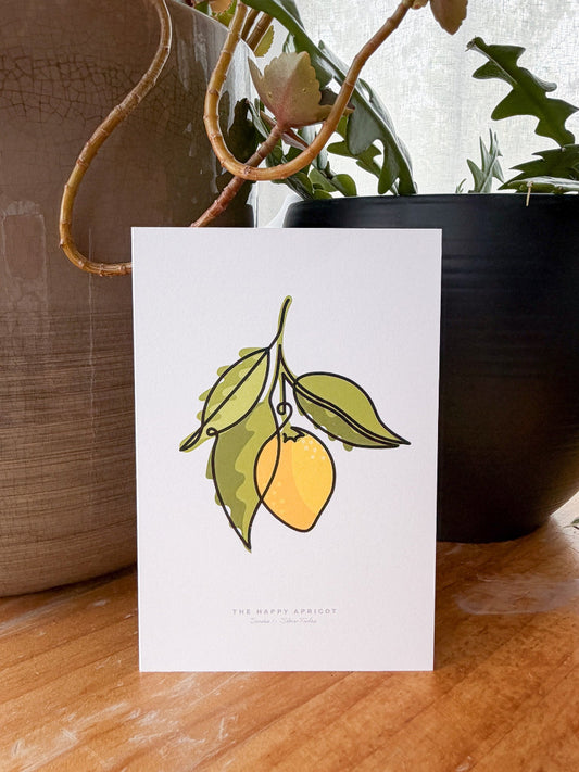 A white greeting card featuring a minimalist illustration of a yellow lemon with green leaves, standing on a wooden surface with potted plants in the background.