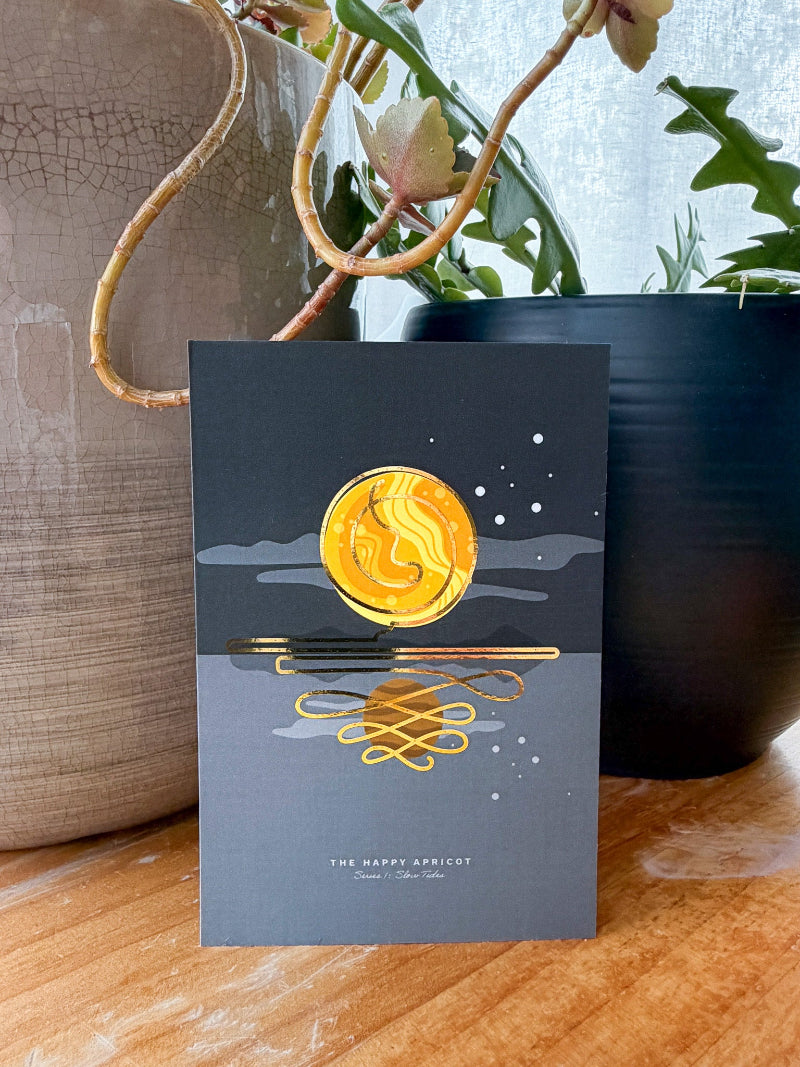 A card featuring a gold crescent moon and reflection design on a dark gray background, styled on a wooden surface with potted plants in the background.