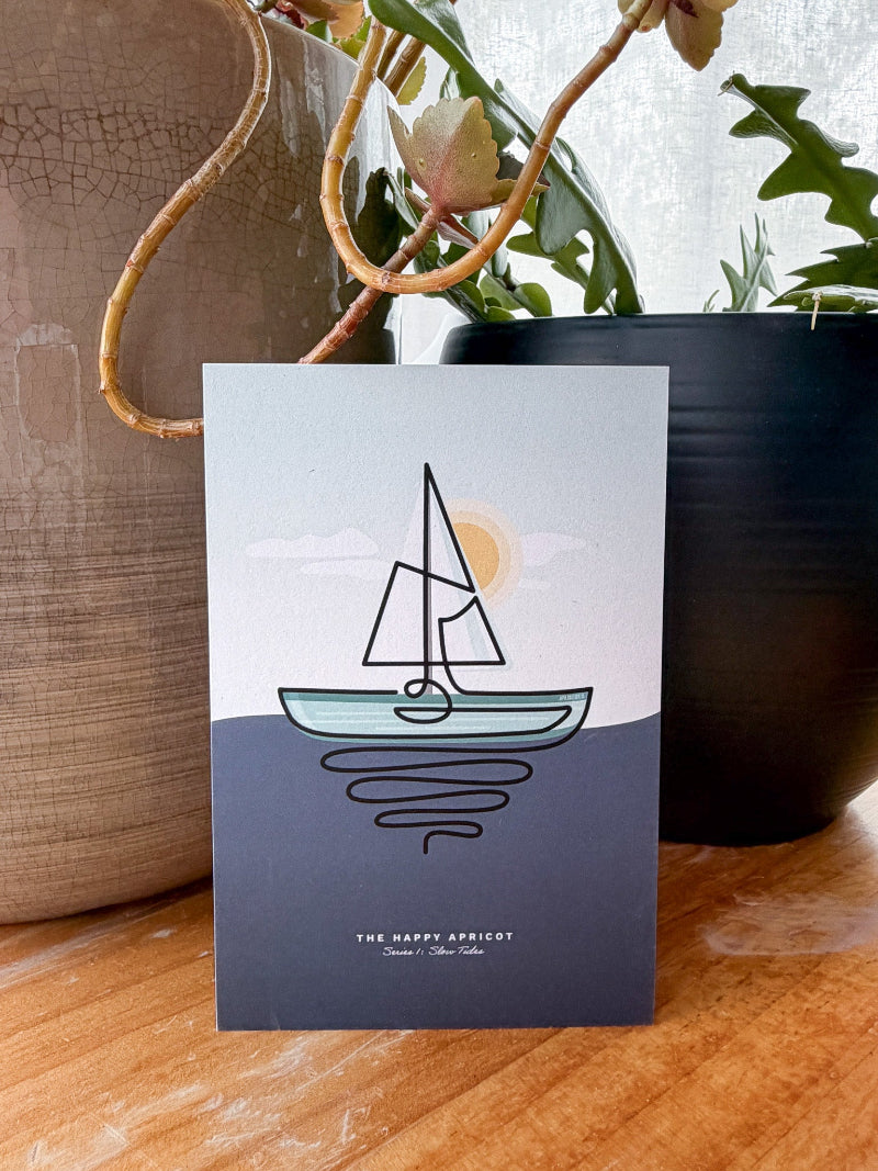 A card featuring a minimalist line art illustration of a sailboat on water, styled on a wooden surface with potted plants in the background.