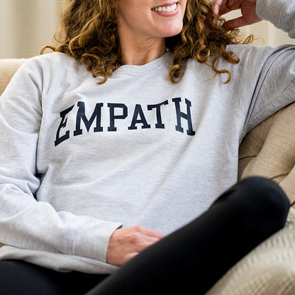 Woman smiling in gray EMPATH crewneck sweatshirt sitting on fabric couch.